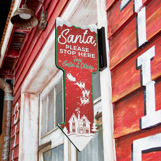 Decorative sign with 'Santa Please Stop Here' text on a red background, attached to a building exterior.