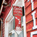 Decorative sign with 'Santa Please Stop Here' text on a red background, attached to a building exterior.