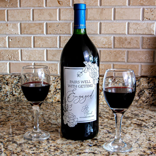 Wine bottle labeled 'Pairs Well with Getting Engaged' with two glasses of red wine on a countertop.