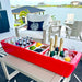 Red outdoor cooler with drinks and snacks on a patio by a waterfront.
