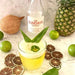 Glass of yellow liquid with green leaves, bottle labeled 'Los Sundays Coconut', pineapple, coconuts, and limes on a marble surface.