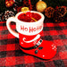 Red mug with 'Ho Ho Ho!' text, filled with a red liquid and a cherry, on a checkered background with Christmas decorations.