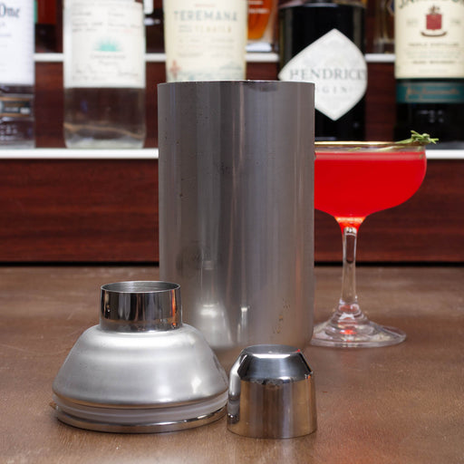 Metal cocktail shaker with two additional components on a bar counter with a red cocktail in the background.