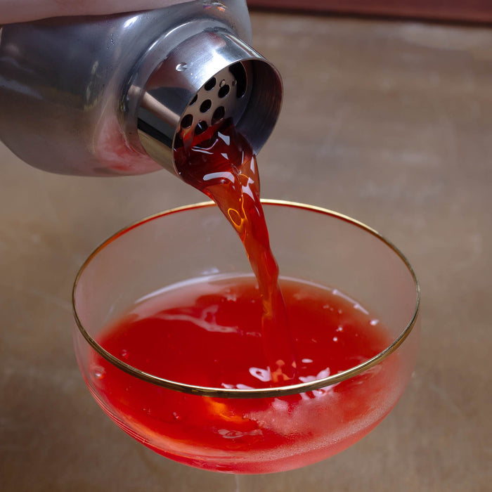 Red liquid being poured from a cocktail shaker into a glass with a gold rim.