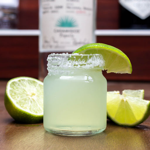 Mini mason jar with a shot of margarita with lime wedge on a wooden surface, with a bottle in the background.