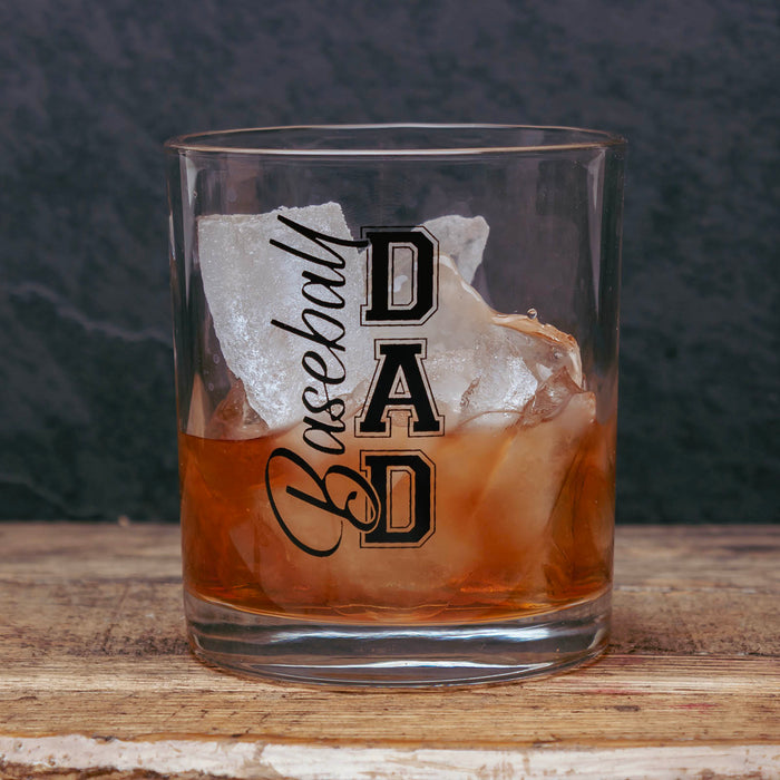 A clear rocks glass with black 'Baseball DAD' lettering, filled with an amber drink and large ice cubes, set on a wooden surface against a dark textured background.