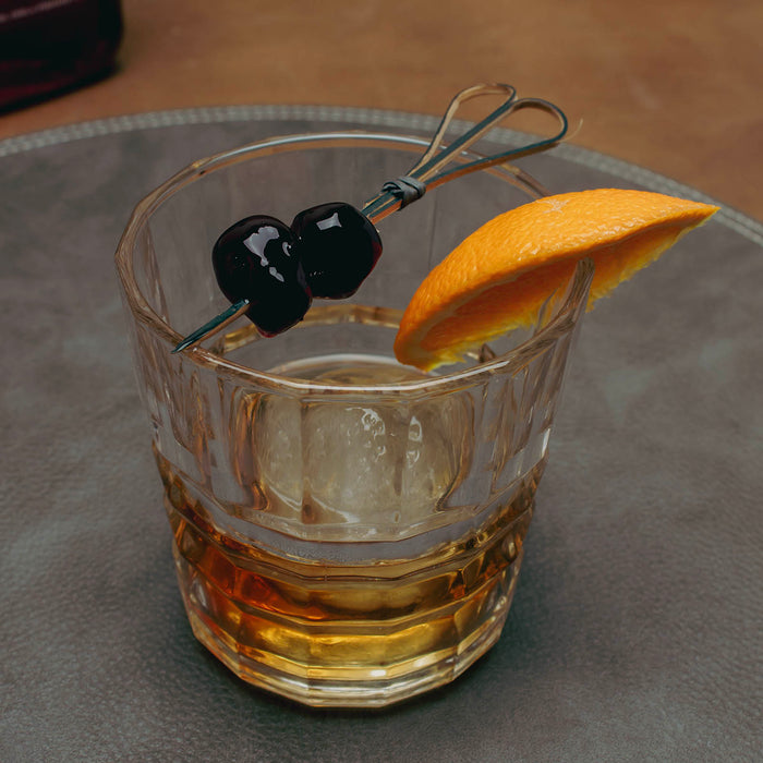 Glass of whiskey with cherries and an orange wedge on a gray surface