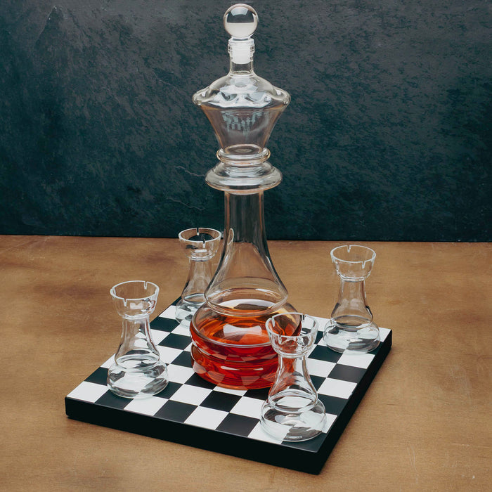 A clear glass decanter with a matching glass stopper and four small clear glass tumblers arranged on a black-and-white checkered board, with the decanter partially filled with amber liquid. The setup sits on a tan surface against a dark textured backdrop.