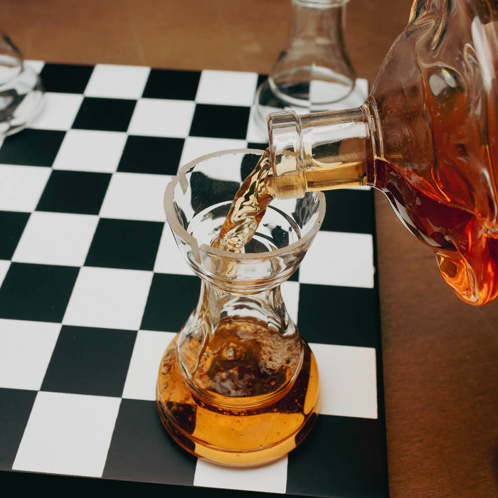 A clear glass decanter pouring amber liquid into a matching clear glass carafe, set on a black-and-white checkered surface with a warm brown background.