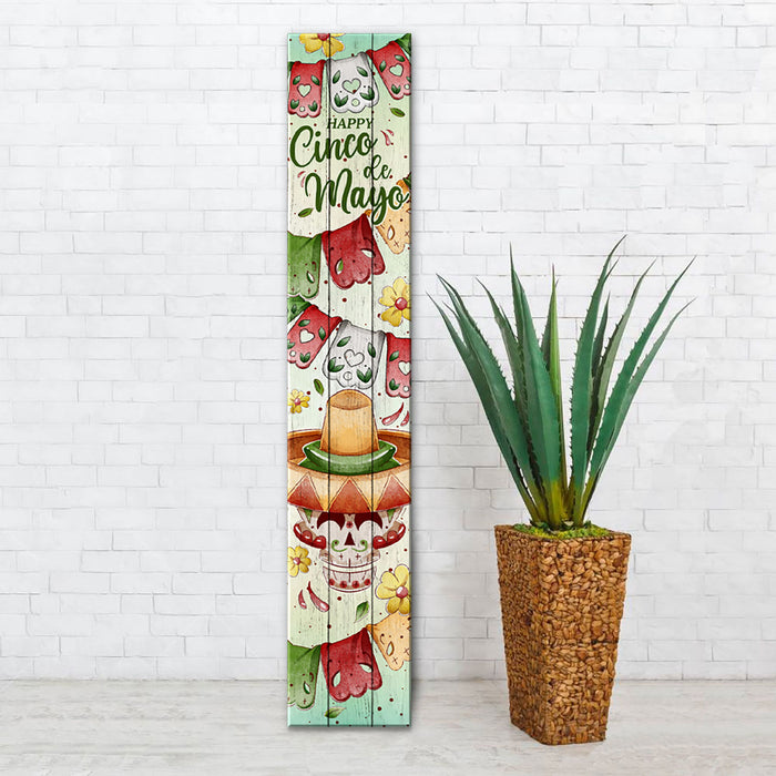 Decorative sign with 'Happy Cinco de Mayo' text next to a potted plant against a white brick wall.