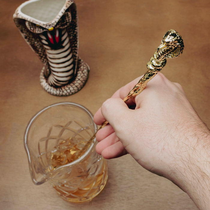 A hand holds a gold-toned metal spoon with a sculpted snake handle above a clear glass beaker containing amber liquid, with a small cobra tiki mug in the background on a tan surface.