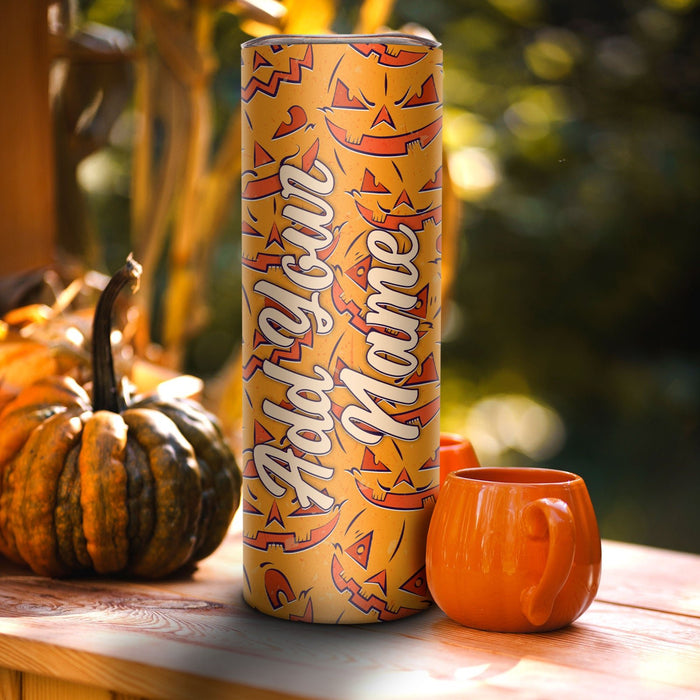 A tall cylindrical mug or tumbler with a Halloween-themed orange and yellow jack-o'-lantern pattern and the words 'Add Your Name' in white script, standing on a wooden table next to an orange mug and pumpkins with a blurred outdoor background.