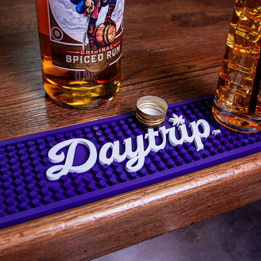 Bottle of Spiced Rum, glass, and purple bar mat with 'Daytrip' branding on a wooden surface.