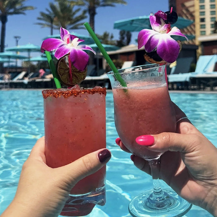 Two pink cocktails with floral garnishes held by hands against a poolside background.