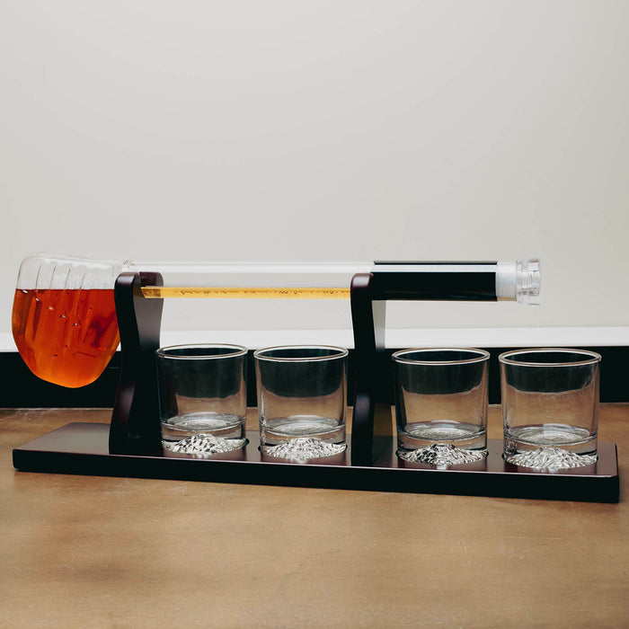A whiskey decanter set on a black base, featuring a clear glass horizontal tube with a black cap and a glass pitcher filled with amber liquid on the left, plus four small clear glasses on the base.