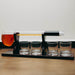 A whiskey decanter set on a black base, featuring a clear glass horizontal tube with a black cap and a glass pitcher filled with amber liquid on the left, plus four small clear glasses on the base.