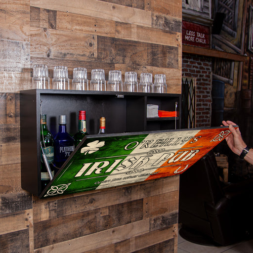 A black wall-mounted bar cabinet with glass holders and bottles, shown in a rustic wood-paneled room. A hand is opening a hinged panel below the cabinet that displays an Irish-themed sign reading 'Irish Pub' with green, white, and orange sections.