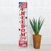 Decorative sign with 'Let Freedom Ring' text and American flag design next to a plant against a white brick wall.