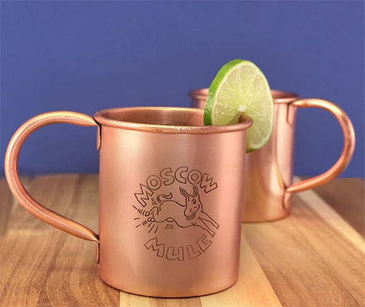 Copper Moscow Mule mugs with lime slices on a wooden surface against a blue background