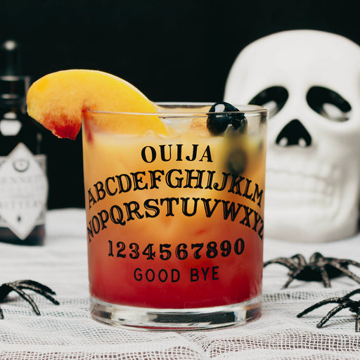 A clear glass tumbler printed with a Ouija board design, filled with a layered orange-to-red cocktail and garnished with a peach slice and black olives. The glass sits on a lace surface with a white skull, black spiders, and a dark bottle in the background.