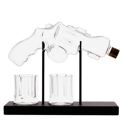 A clear glass revolver-shaped bottle holder on a black base with two small clear glasses, photographed against a plain white background.