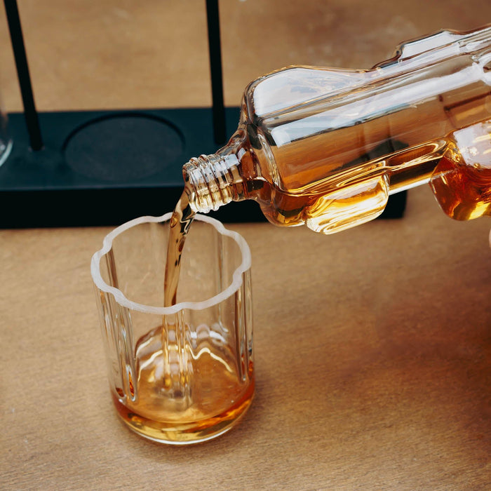 A clear glass bottle with a ribbed neck and scalloped rim pours amber-colored liquid into a matching clear ribbed glass tumbler on a warm wood surface.