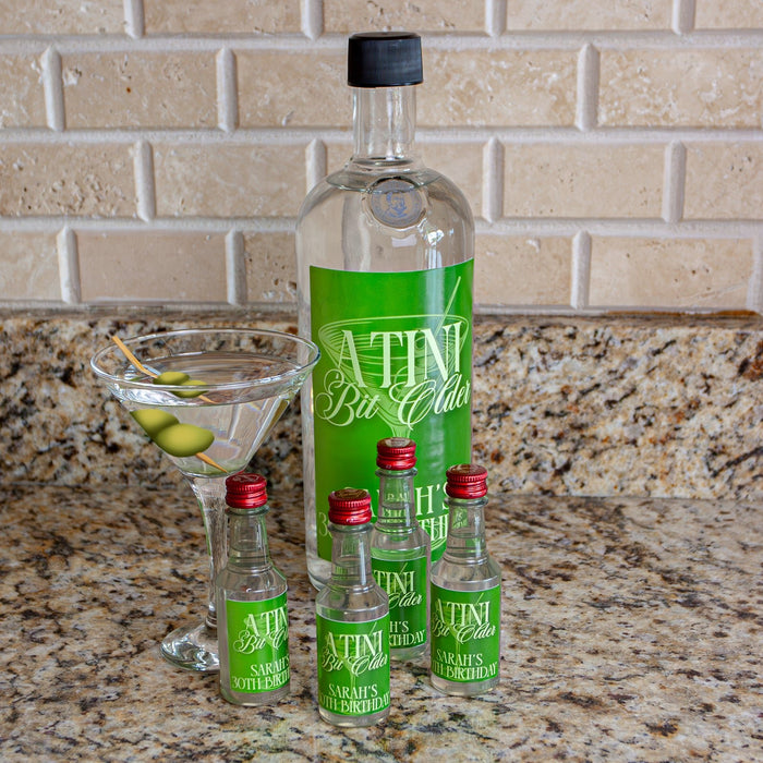 Bottle with mini bottles and a cocktail on a countertop with 'A Tini Bit Older' stickers.