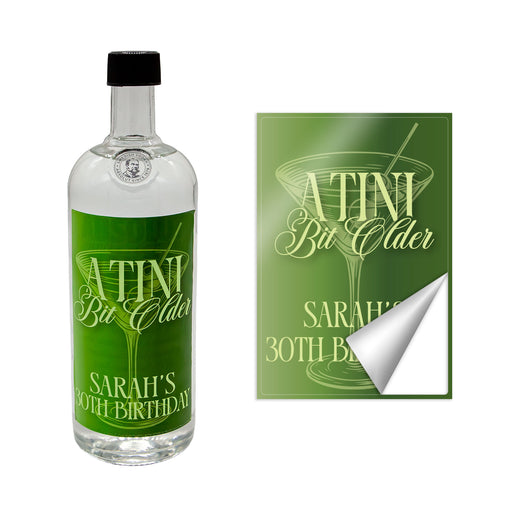 Bottle with a customizable 'A Tini Bit Older' green label and a matching sticker on a white background
