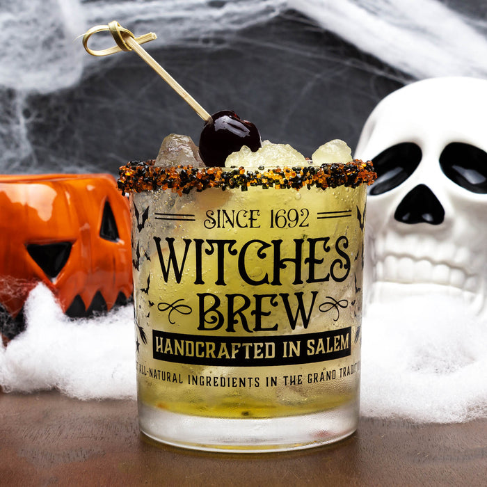 Cocktail in a glass labeled 'Witches Brew' with Halloween decorations in the background.